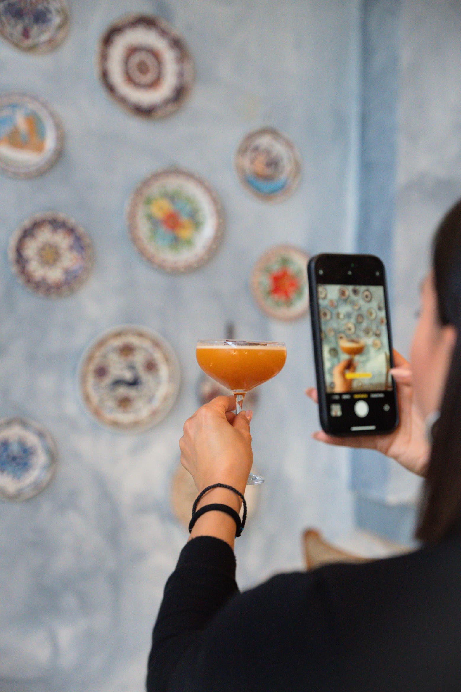 A guest photographing her drink by a whitewashed wall of traditional plates